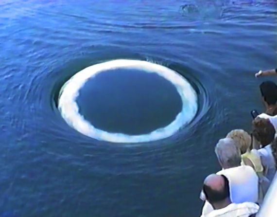 video still of people on a boat looking at w white ring on the ocean